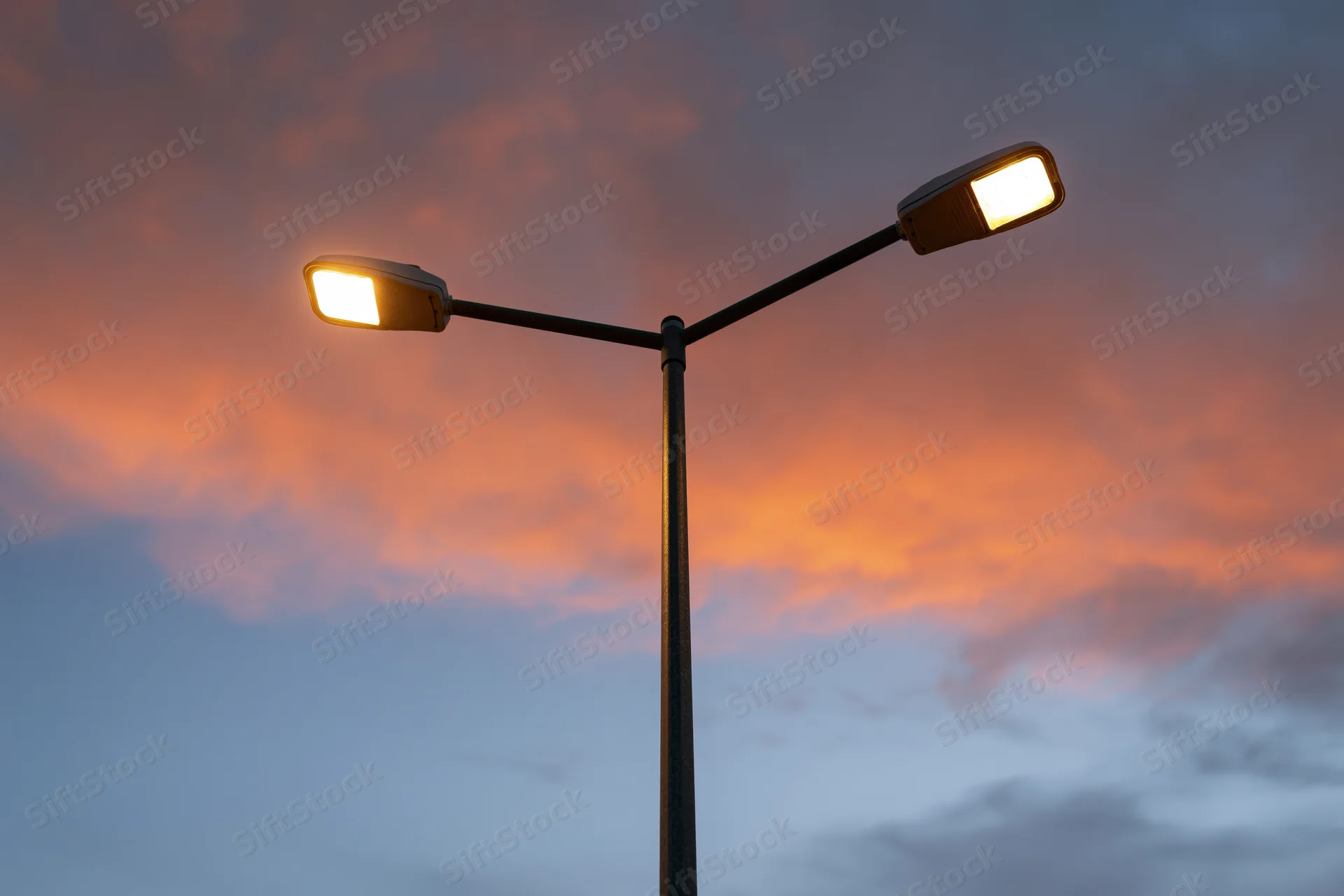 Dual-arm led street lamp pole illuminated against twilight sky with orange and pink clouds