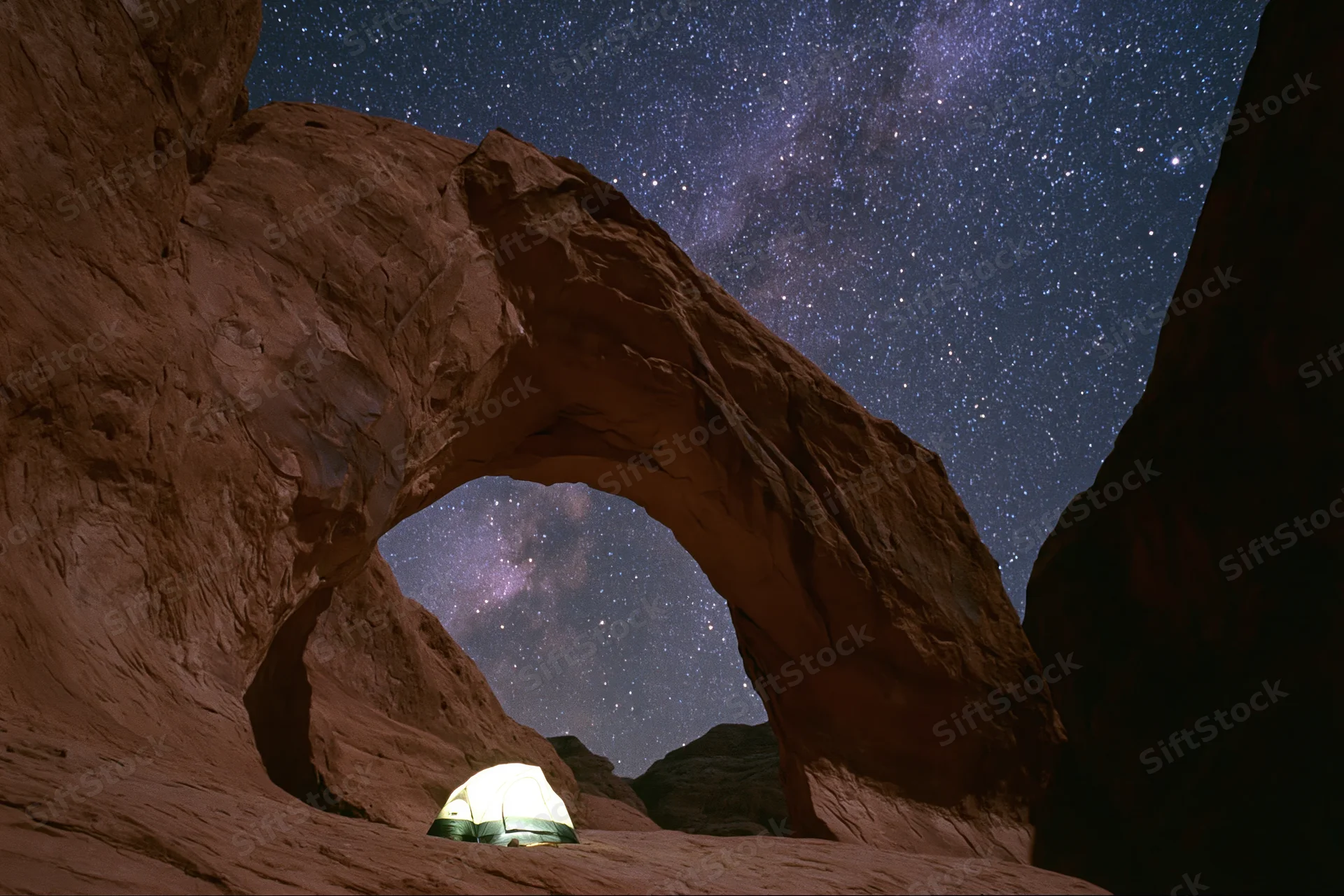Glowing tent beneath massive sandstone arch under starry night sky in desert landscape