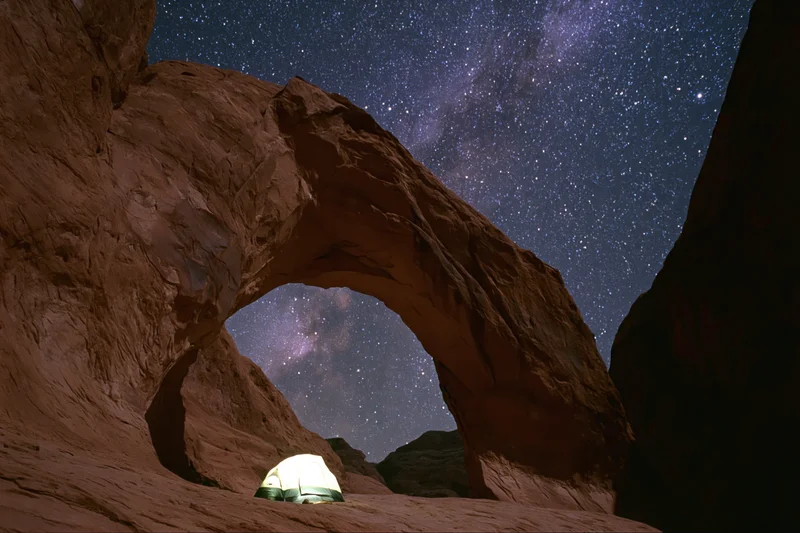 Glowing tent beneath massive sandstone arch under starry night sky in desert landscape