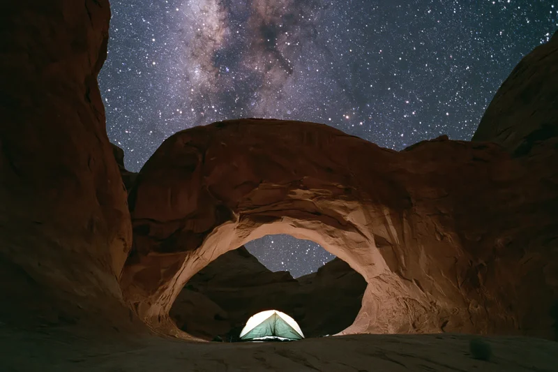 Glowing tent beneath massive sandstone arch in desert at night under starry sky