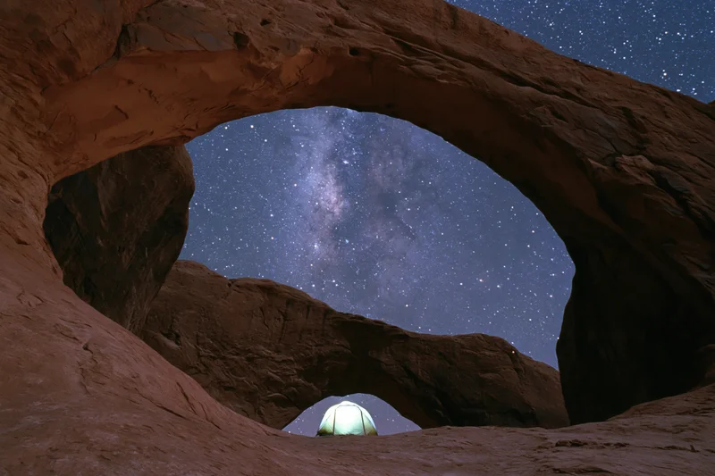 Glowing tent beneath massive sandstone arch under starry night sky in desert wilderness