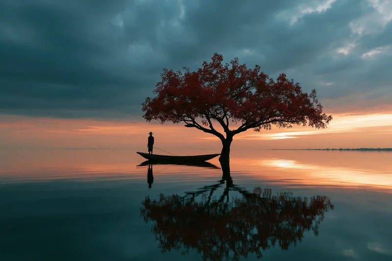 Fisherman standing in a small wooden boat beside a red-leafed tree in calm water at sunset