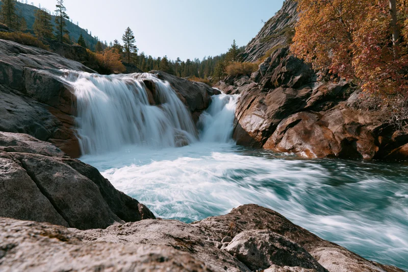 Waterfall cascading over granite ledges into a rushing river surrounded by autumn trees and mountain slopes