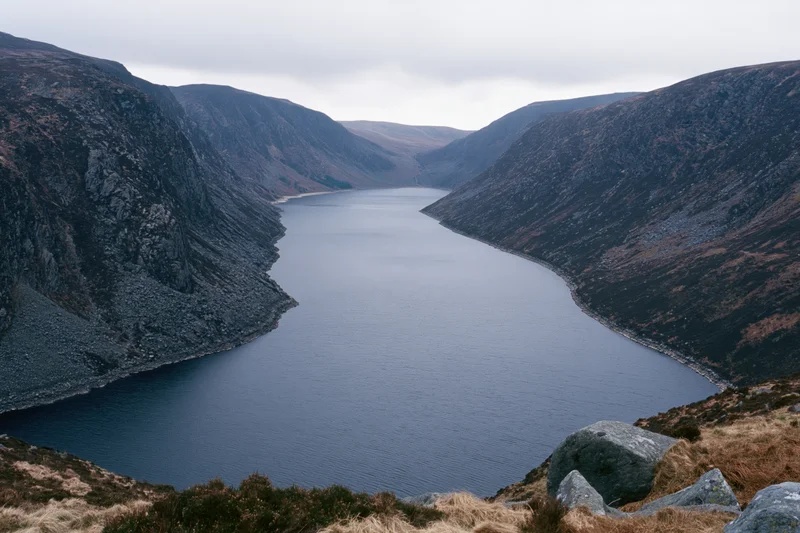 Narrow mountain reservoir stretching into deep valley surrounded by rocky slopes under overcast sky