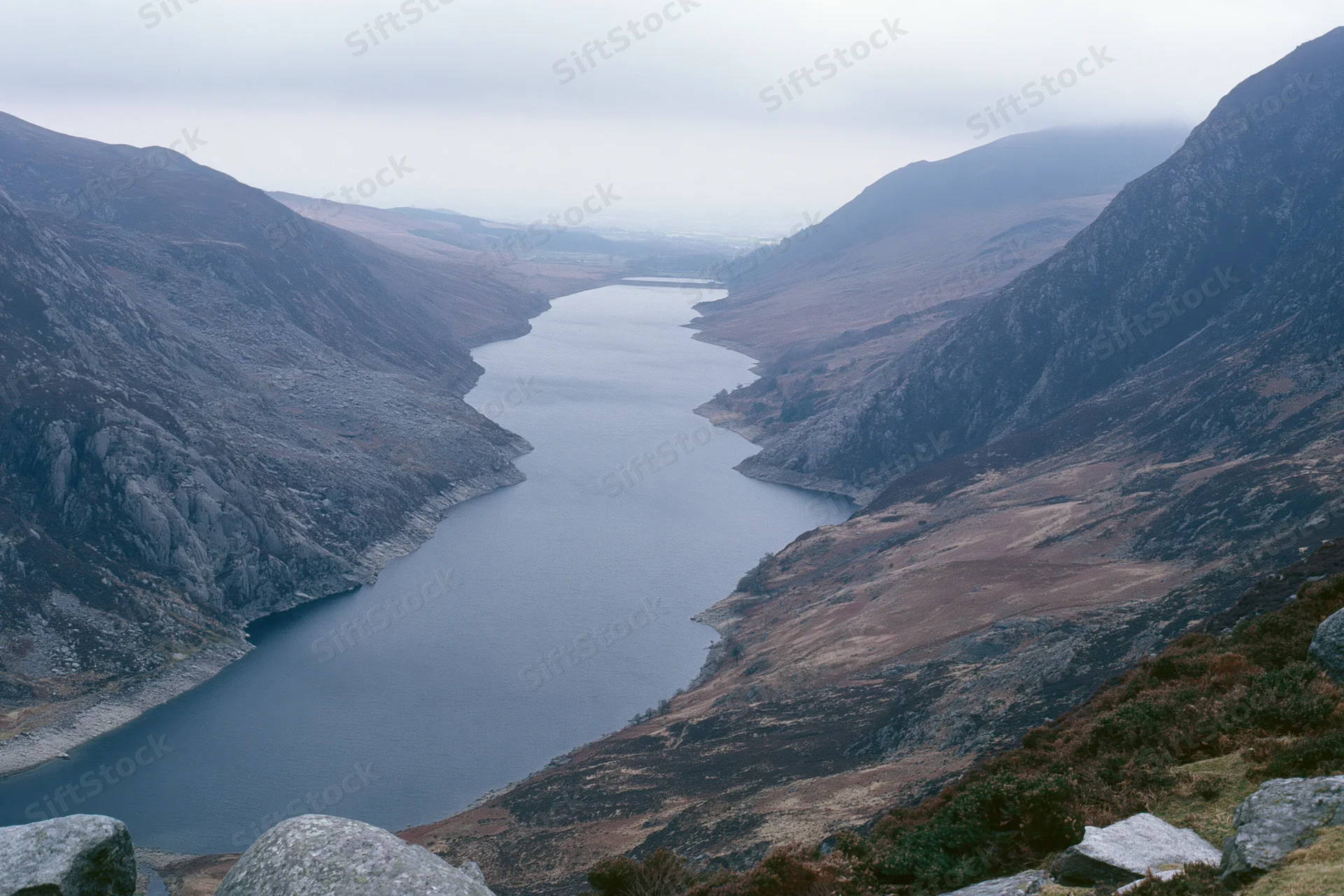 Narrow mountain reservoir stretching into deep valley surrounded by rocky slopes under overcast sky
