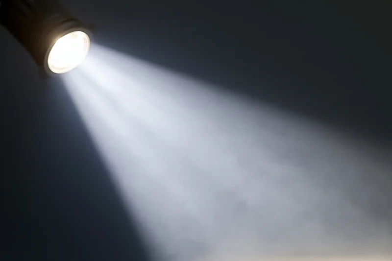 Single spotlight mounted in upper left casting a sharp beam of white light into dark space