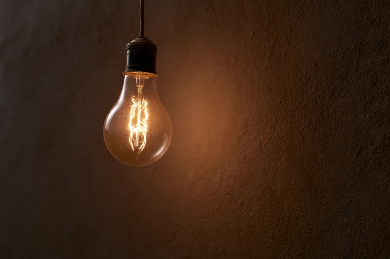 Vintage incandescent lightbulb hanging from ceiling against dark textured wall with warm glow