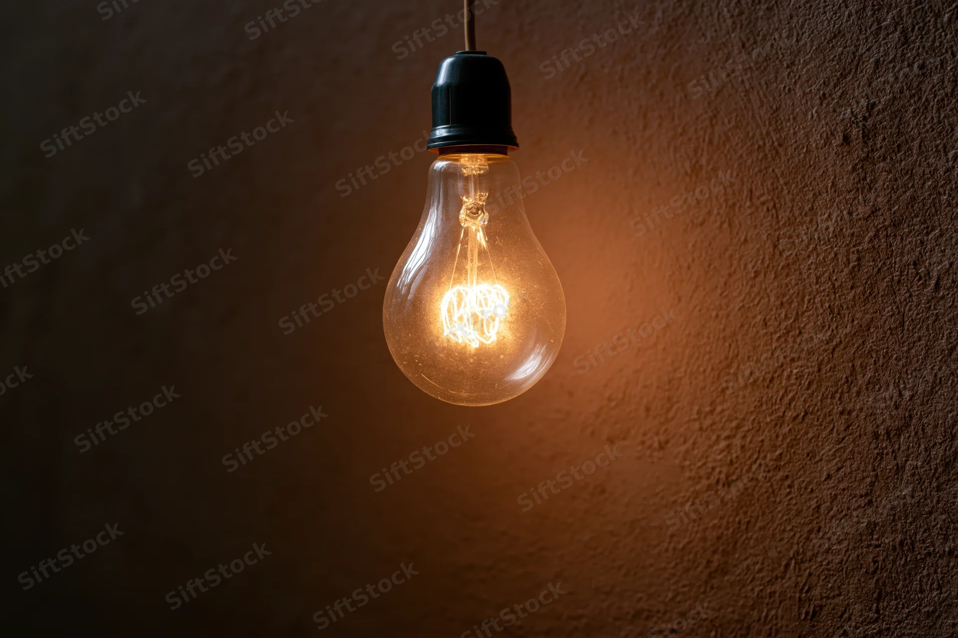 Vintage incandescent lightbulb glowing warmly while hanging from a thin wire against a textured brown wall