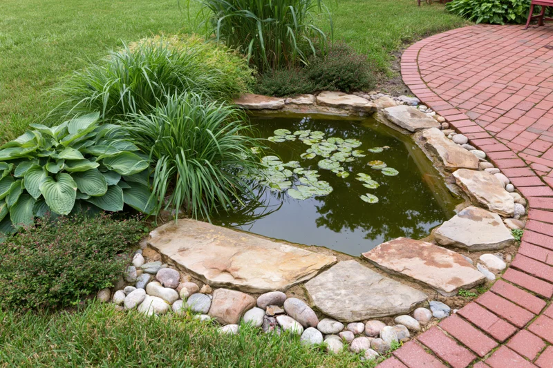 Small garden pond edged with flat sandstone slabs surrounded by green plants and a brick path