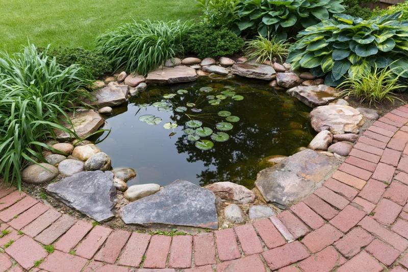 Small garden pond edged with flat sandstone slabs and surrounded by green plants and a brick pathway