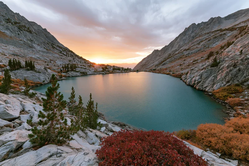 Turquoise alpine lake nestled in rocky mountain valley at sunset with autumn foliage and pine trees surrounding it