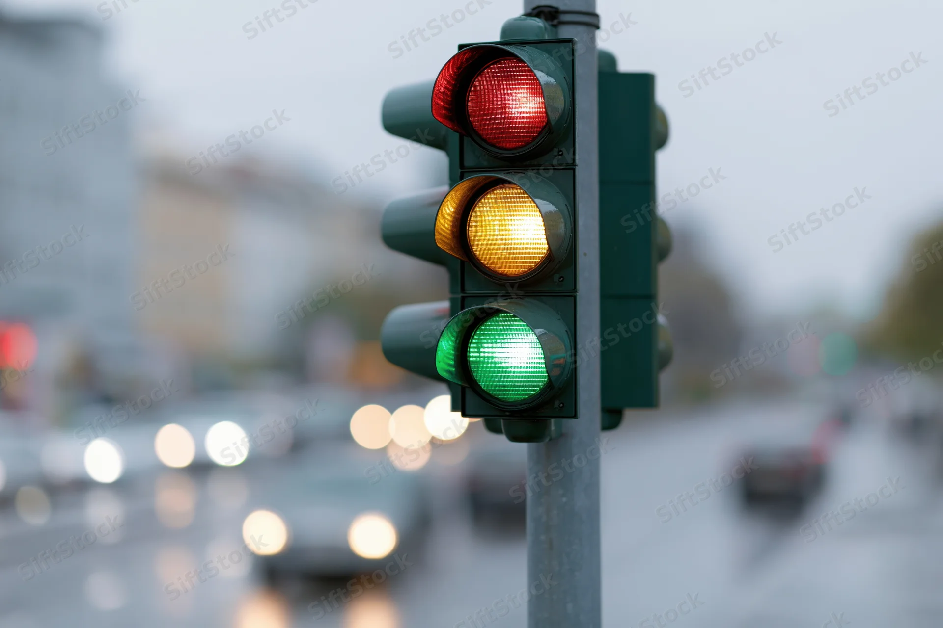 Traffic light showing green at bottom and yellow in middle on city street with blurred vehicles and wet pavement