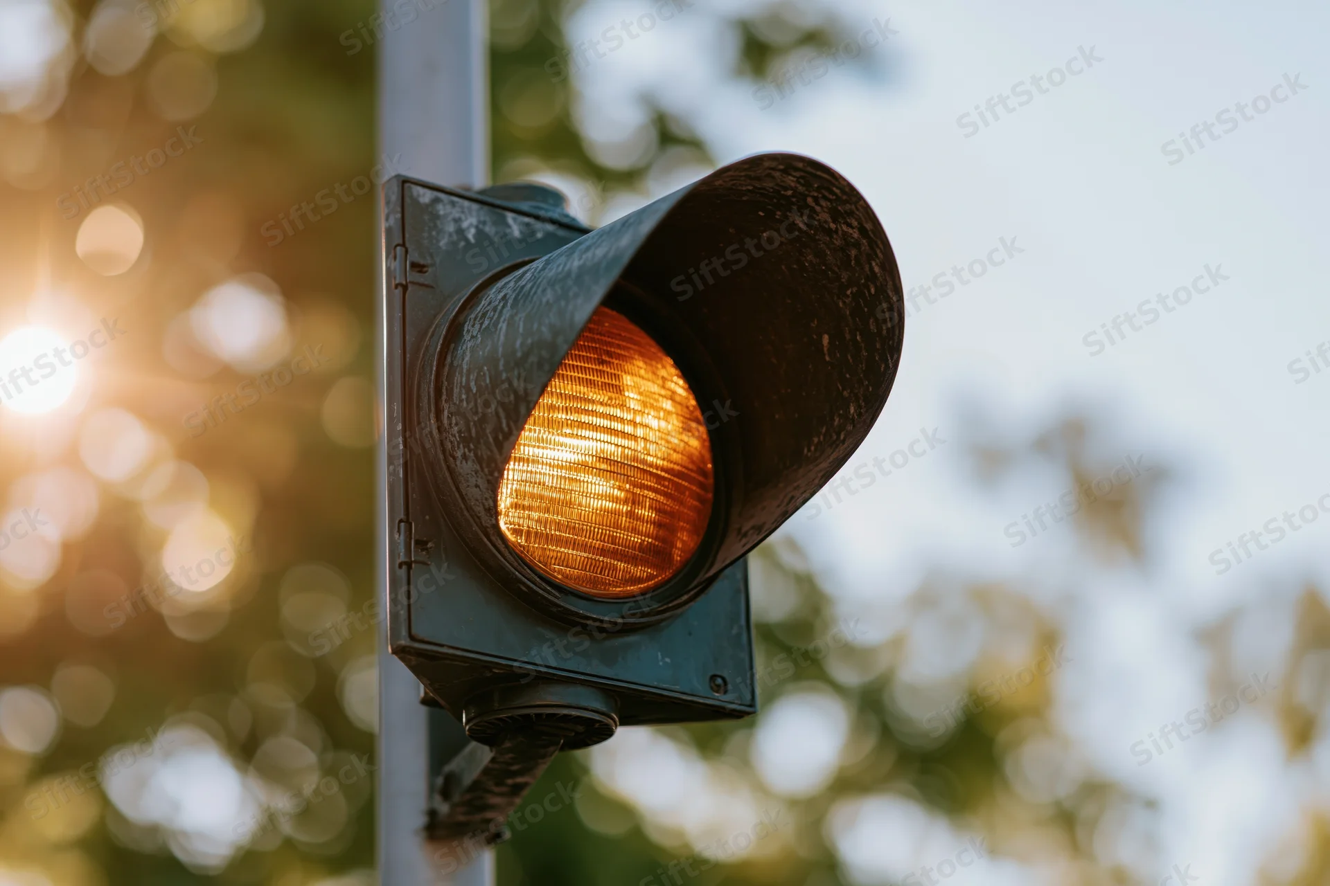 Weathered black metal traffic light with amber signal illuminated on a street pole during daytime with blurred foliage and sunlight in the background