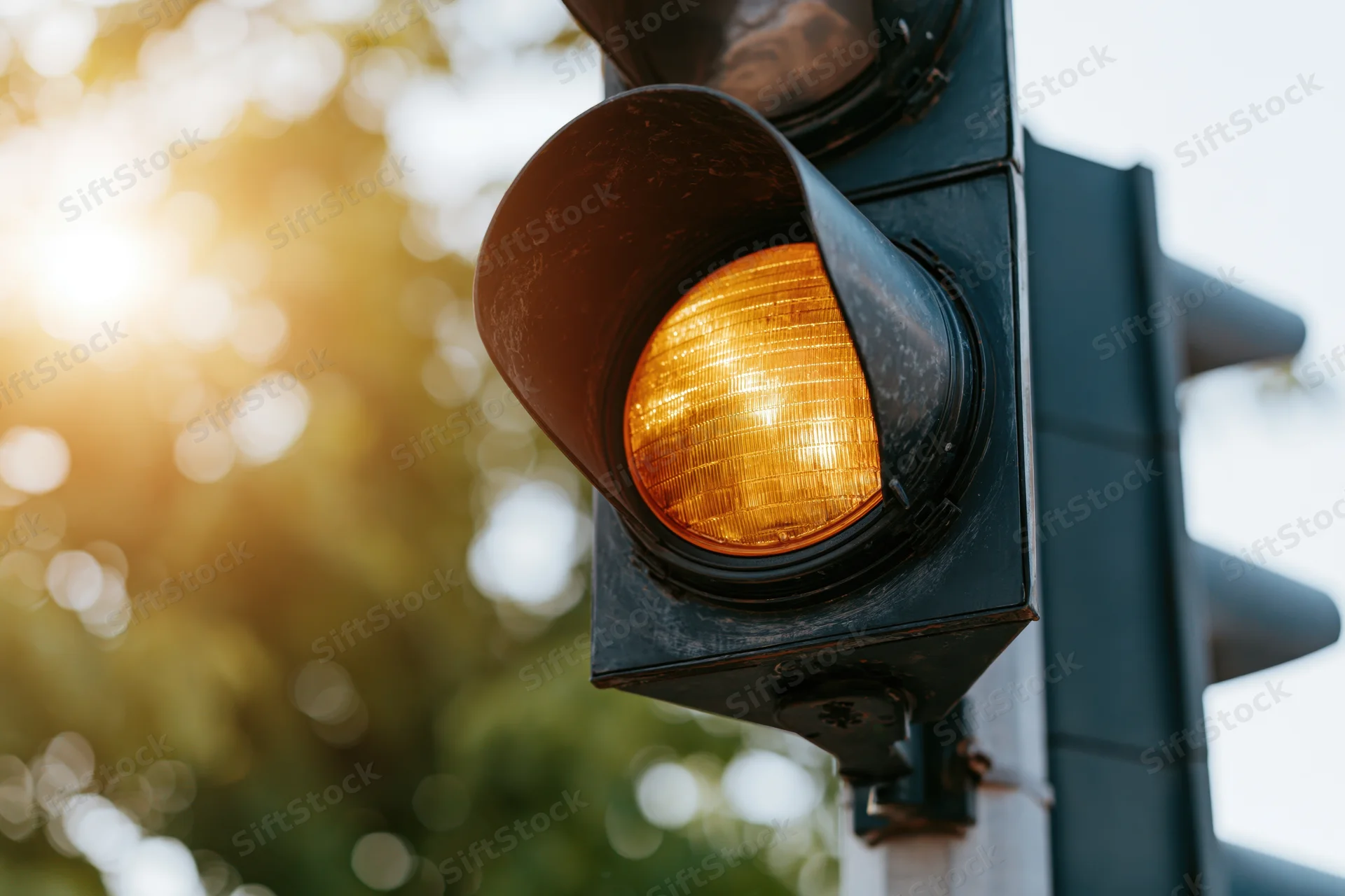 Weathered black metal traffic light with amber signal illuminated against blurred greenery and sunlight