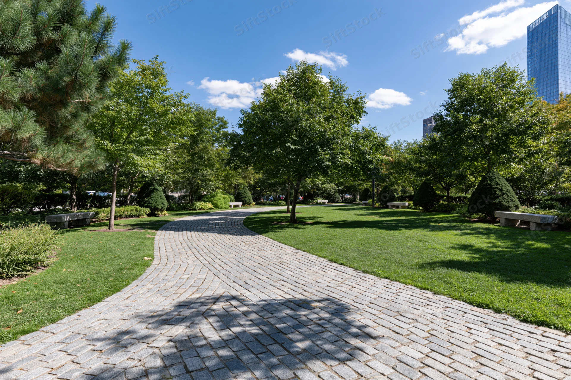 Winding cobblestone path through manicured urban park with trees and benches under blue sky
