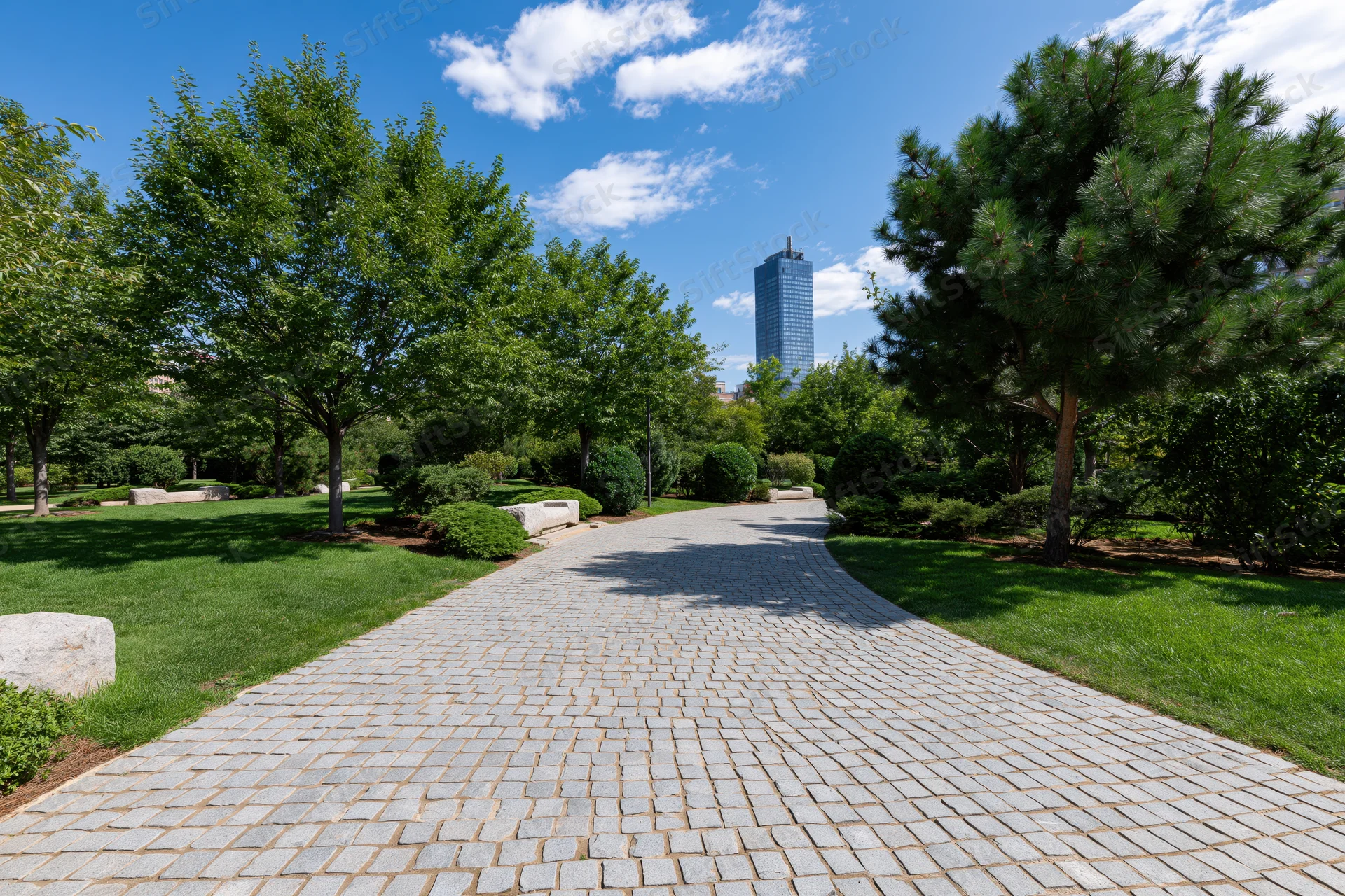 Winding cobblestone path through manicured urban park with trees and distant skyscraper under blue sky