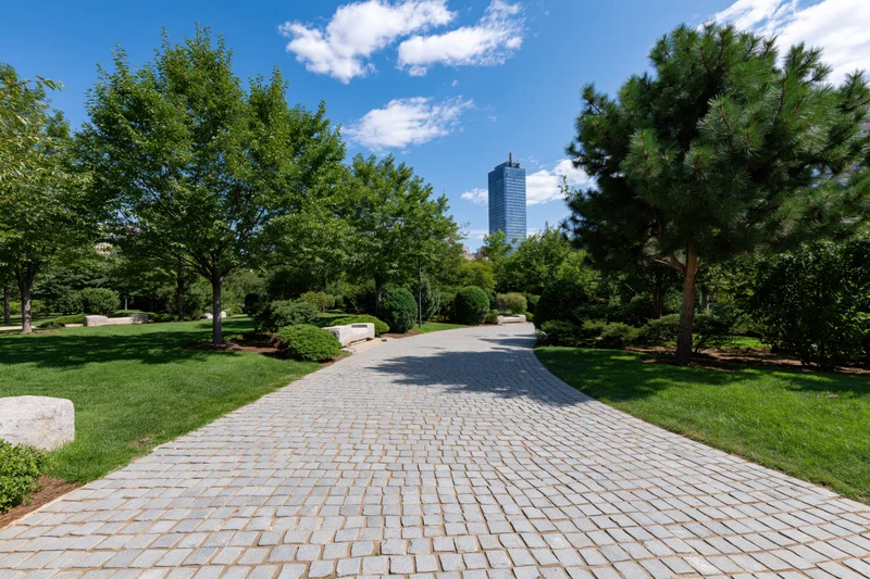 Winding cobblestone path through manicured urban park with trees and distant skyscraper under blue sky