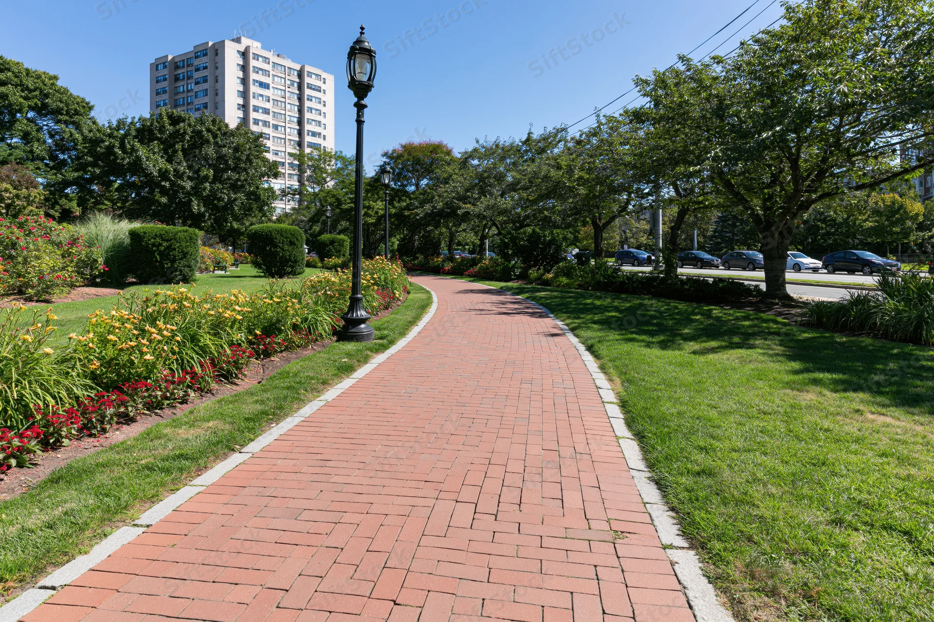 Winding red brick pedestrian path curves through manicured park with trees, flowers, and lampposts, bordered by lawn and urban buildings in background
