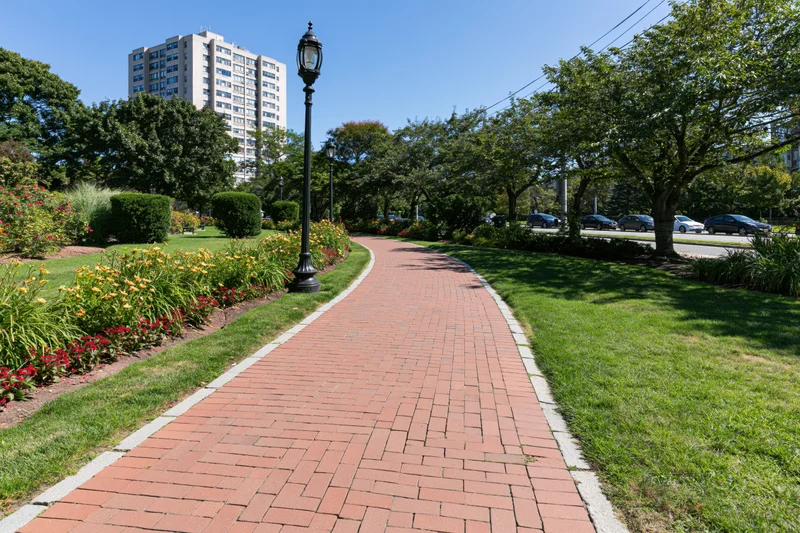 Winding red brick pedestrian path curves through manicured park with trees, flowers, and lampposts, bordered by lawn and urban buildings in background