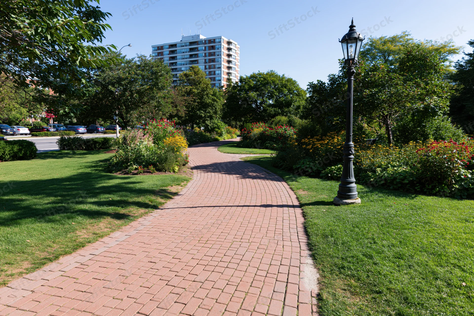 Winding red brick pathway through manicured park garden with trees and flowers near urban apartment building