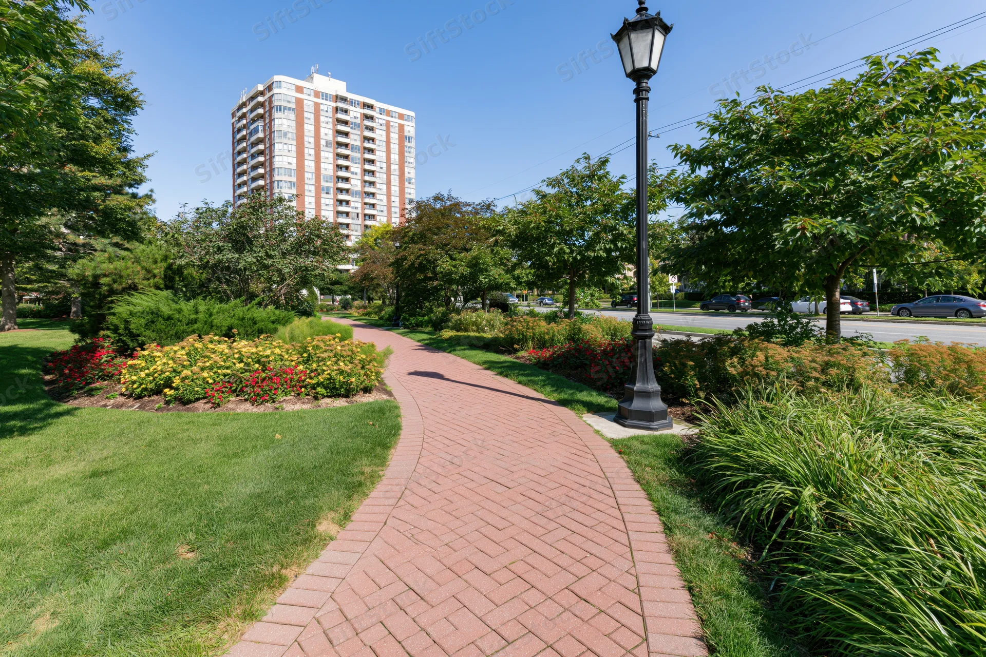 Winding red brick pedestrian path curves through manicured garden with trees and flowers near urban apartment building