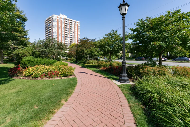 Winding red brick pedestrian path curves through manicured garden with trees and flowers near urban apartment building