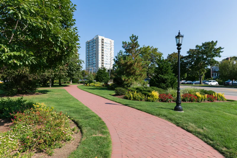 Winding red brick pathway curves through manicured park garden with trees and flowerbeds near urban highrise building