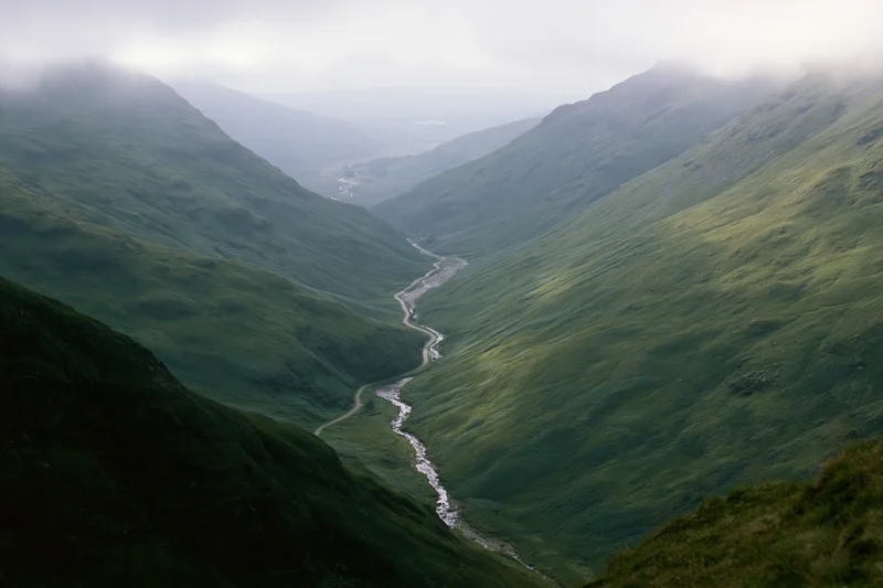 Winding single-track road and narrow stream running through a green mountain valley in the Scottish Highlands