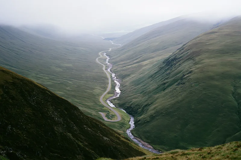 Winding single-track road and narrow stream running parallel through a green mountain valley