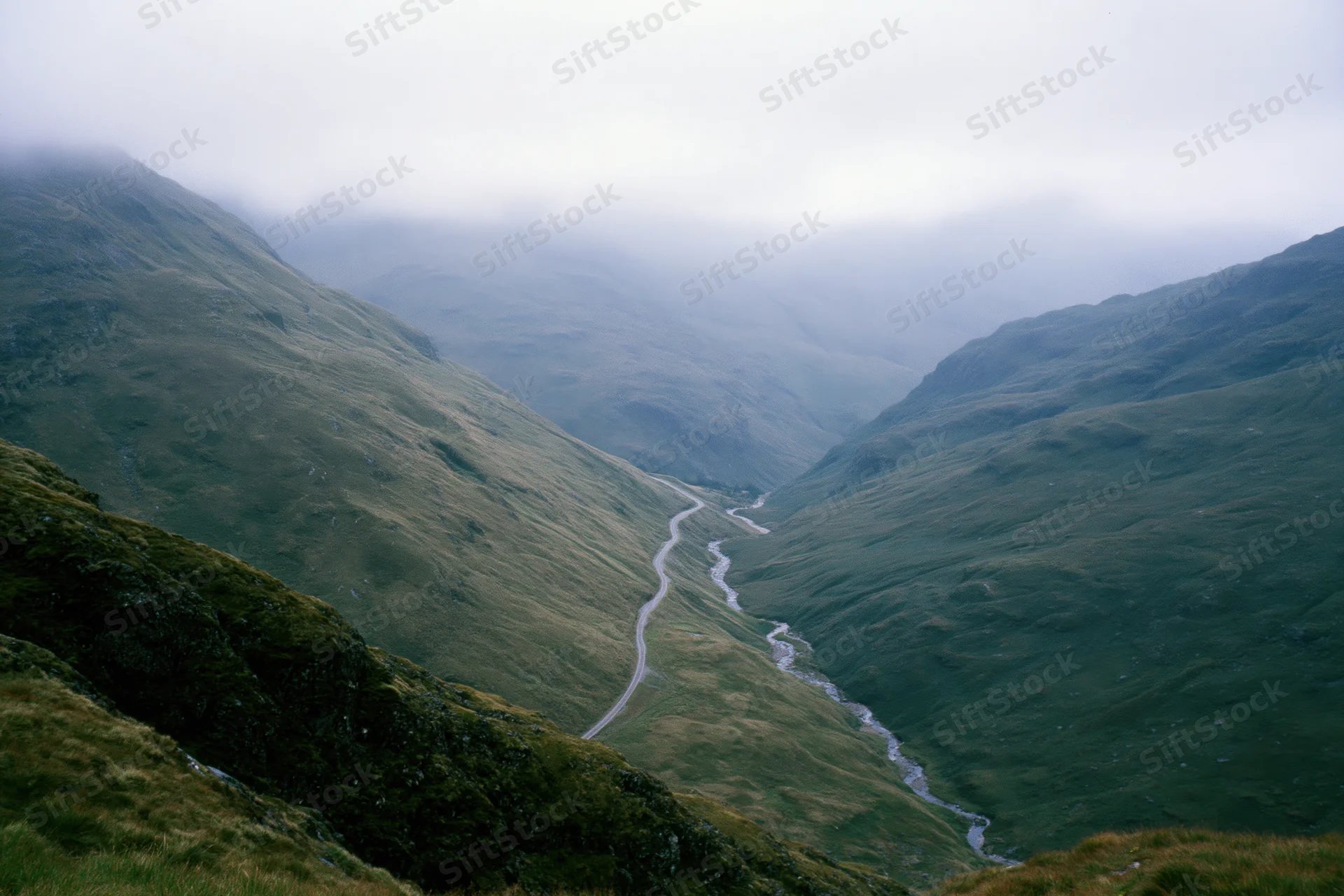 Winding single-track road and narrow stream running parallel through a misty mountain valley