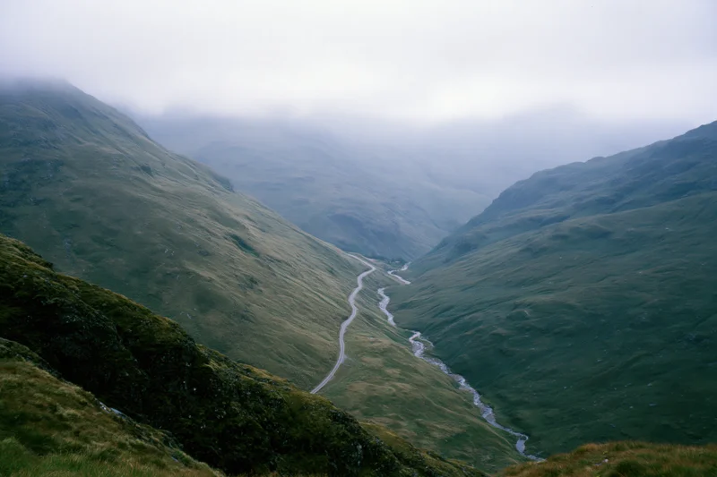 Winding single-track road and narrow stream running parallel through a misty mountain valley