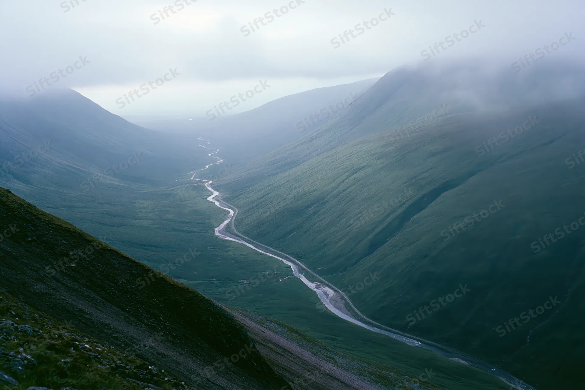 Winding single-track road and narrow stream running through a misty green valley between mountains