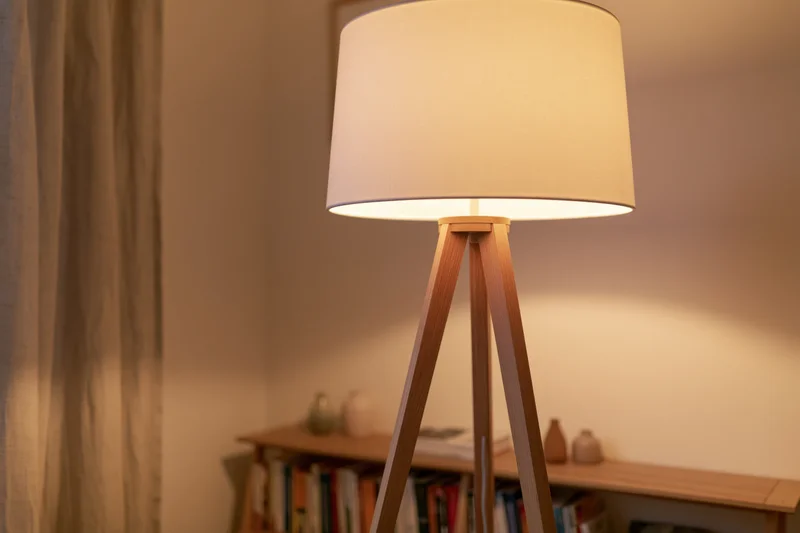 Wooden tripod floor lamp with white drum shade glowing warmly in a living room interior near a bookshelf