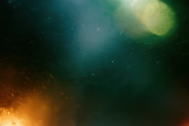 Abstract close-up of a dusty scratched glass surface with soft bokeh lights and scattered particles in dark teal and warm orange tones