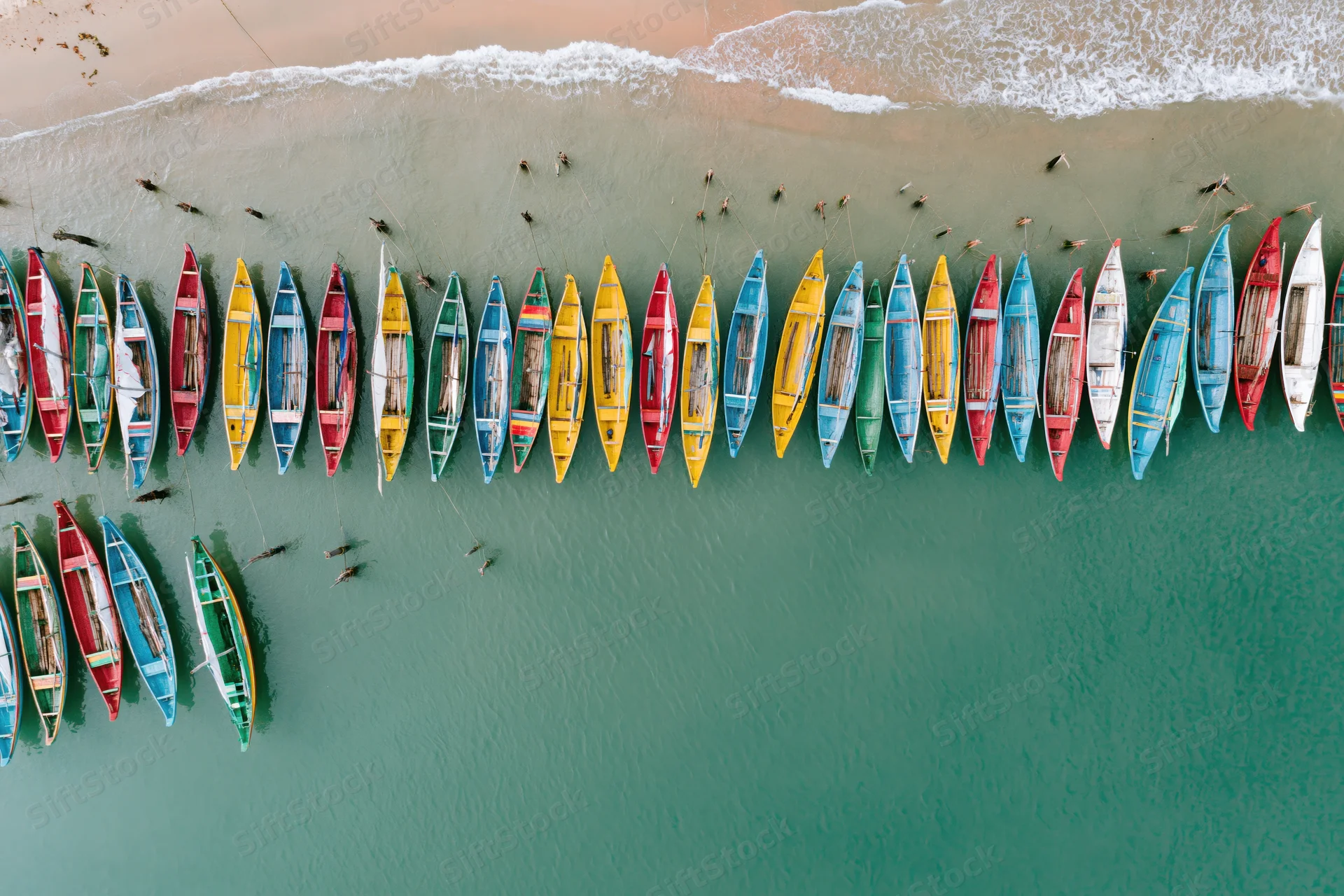 Colorful traditional fishing boats moored in shallow water along a sandy beach from an aerial top-down view