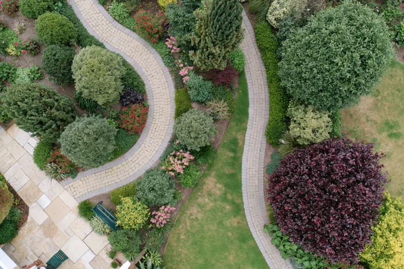 Aerial top-down view of a manicured residential garden with winding stone pathways, shrubs, trees, and flowering plants arranged in landscaped beds