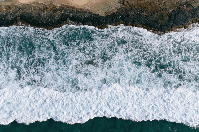Ocean waves breaking over rocky coastline from aerial top-down view