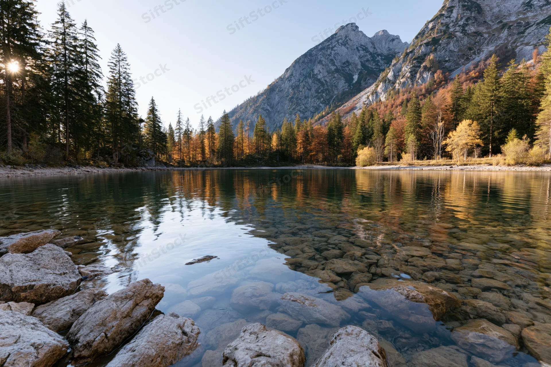 Alpine lake with clear shallow water and submerged rocks surrounded by autumn forest and mountains
