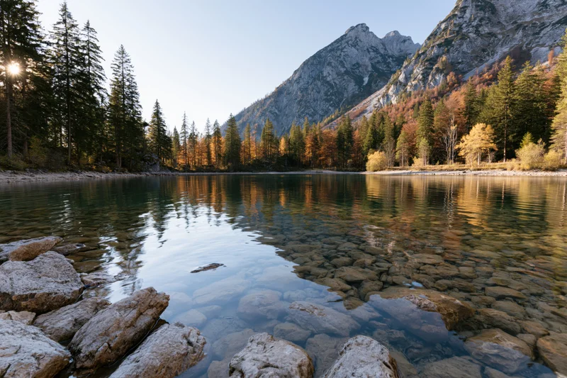 Alpine lake with clear shallow water and submerged rocks surrounded by autumn forest and mountains