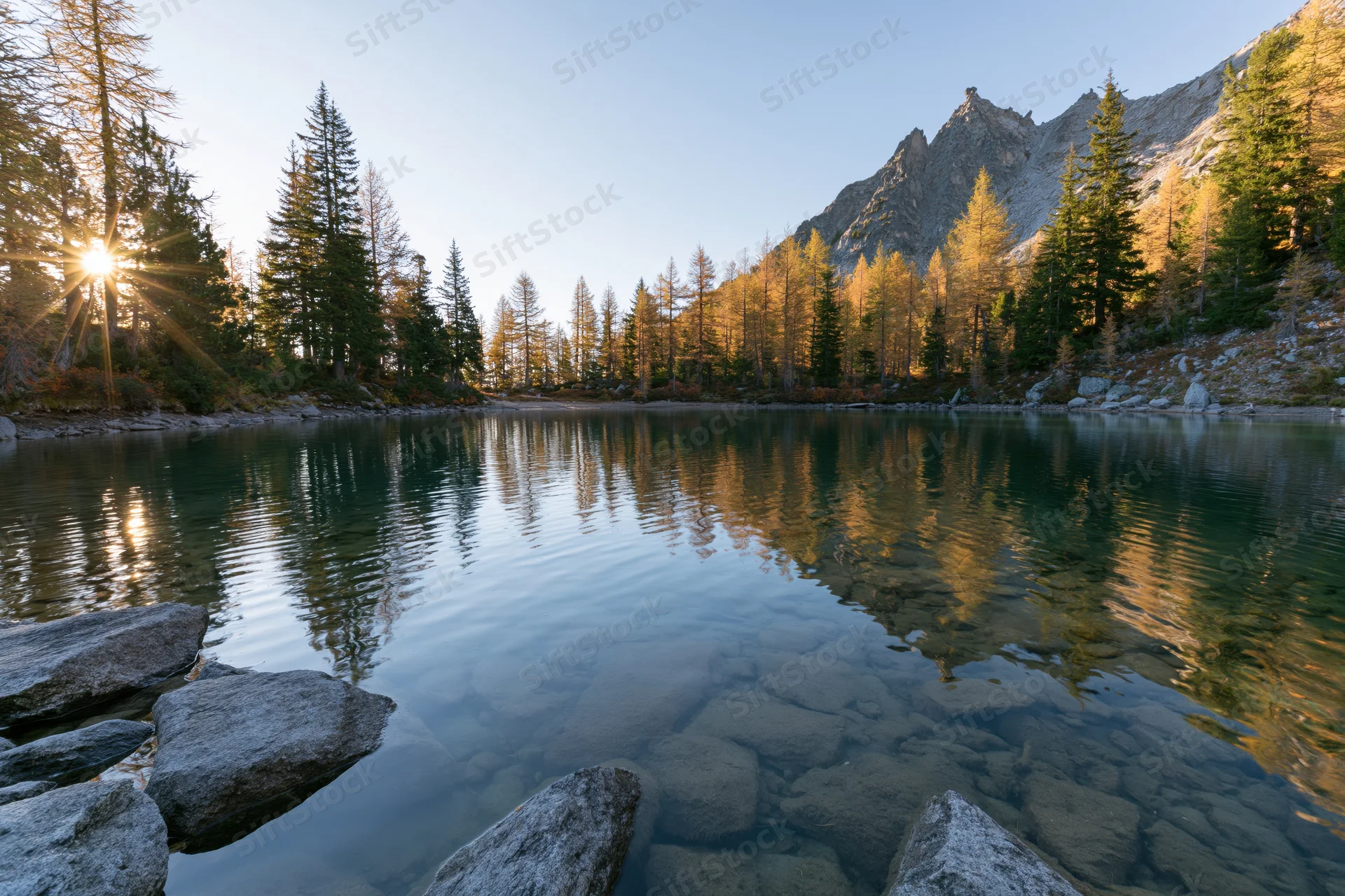 Alpine lake with clear shallow water and submerged rocks reflecting autumn forest and mountain at sunrise