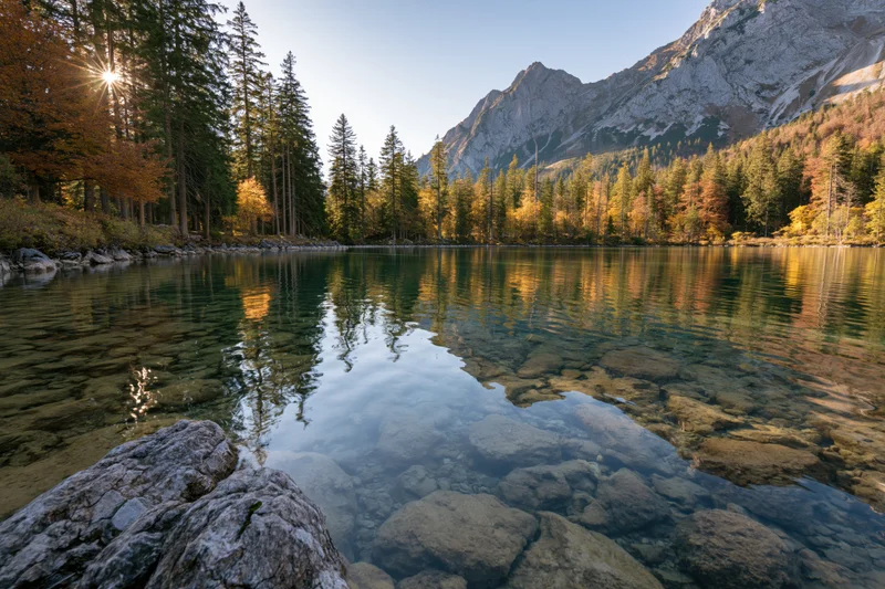 Alpine lake with clear shallow water and submerged rocks surrounded by autumn forest and mountains