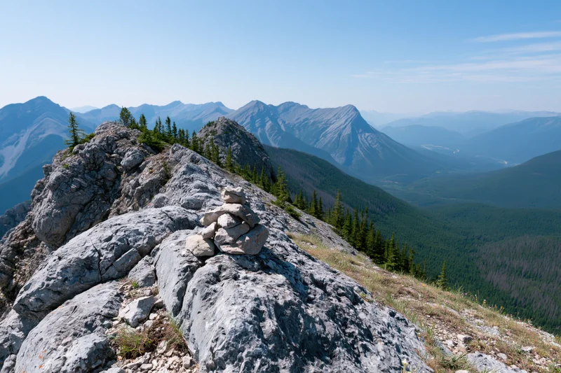 Small stone cairn on rocky alpine ridge overlooking forested valley and distant mountains