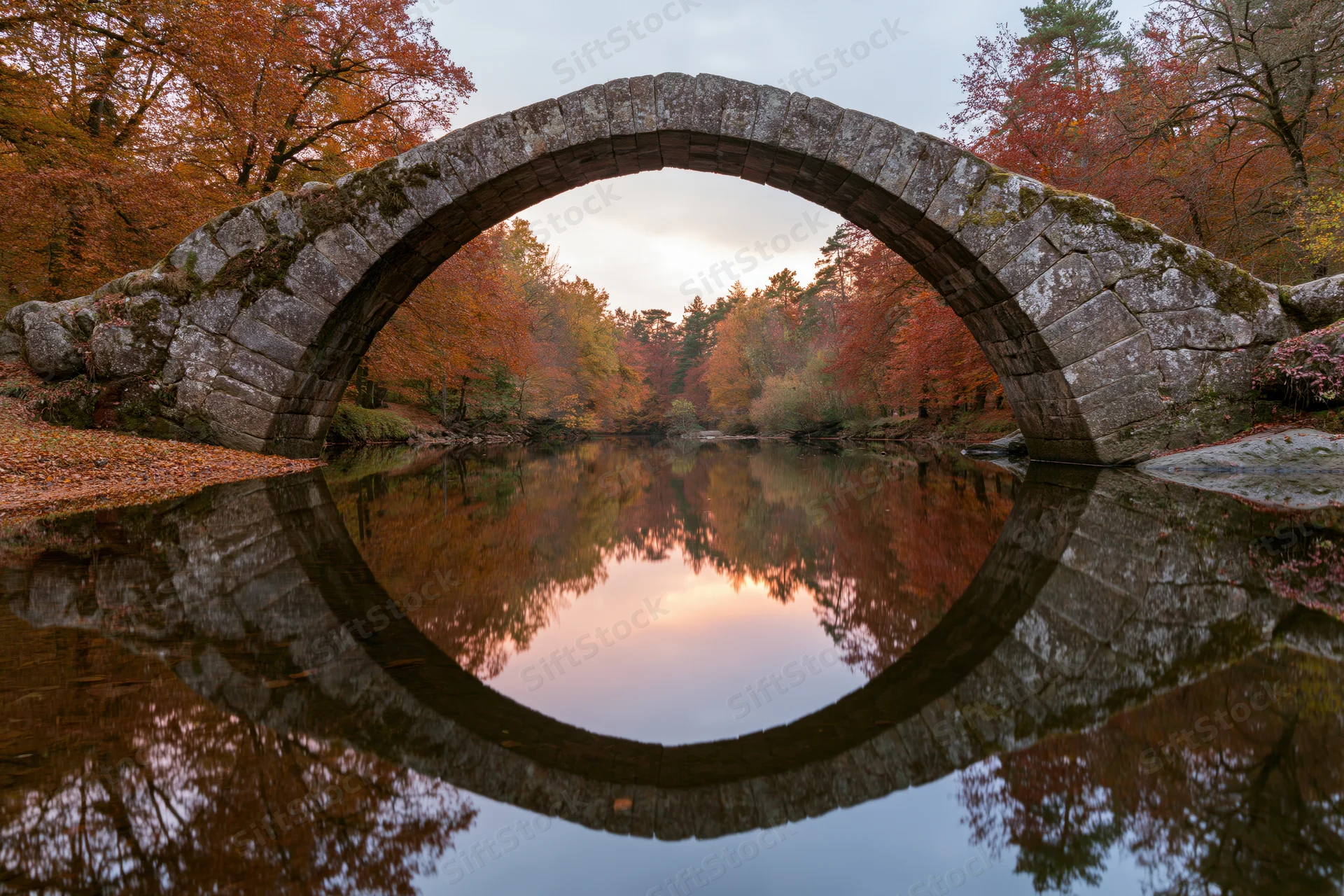 Ancient stone arch bridge spanning a still river with autumn foliage reflected in the water