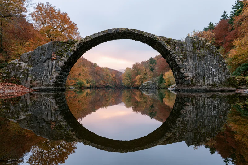 Ancient stone arch bridge spanning a still river with perfect reflection in autumn foliage
