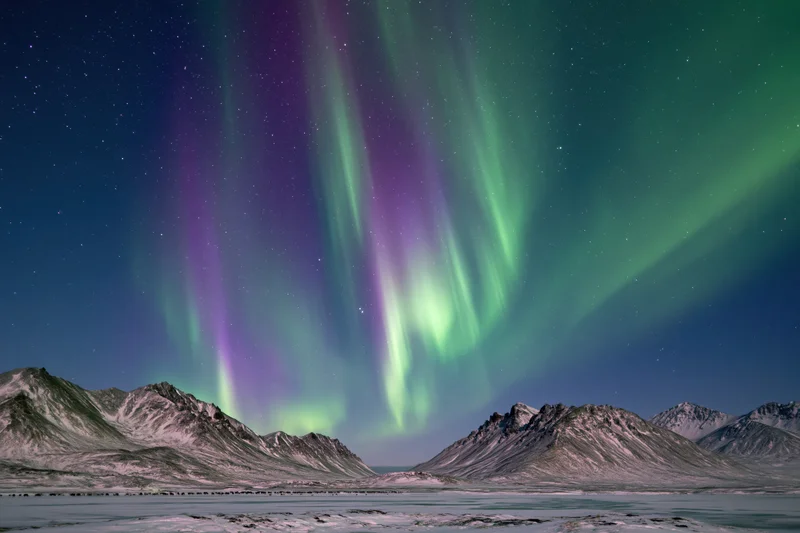 Aurora borealis glowing over snow-covered mountains and frozen lake at night