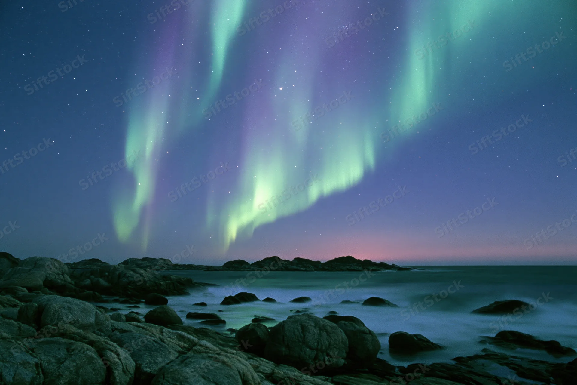 Aurora borealis sweeping in green and purple curtains over rocky coastline at night