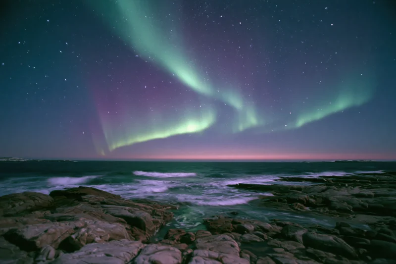 Aurora borealis sweeping across night sky over rocky coastline with ocean waves