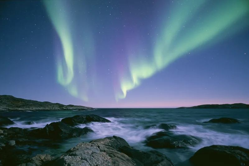 Aurora borealis sweeping in green and purple curtains over rocky coastline at night