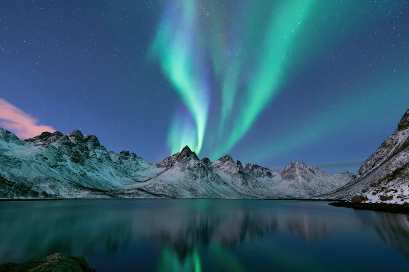 Aurora borealis sweeping in green and blue curtains over snow-covered mountains reflected in calm lake water
