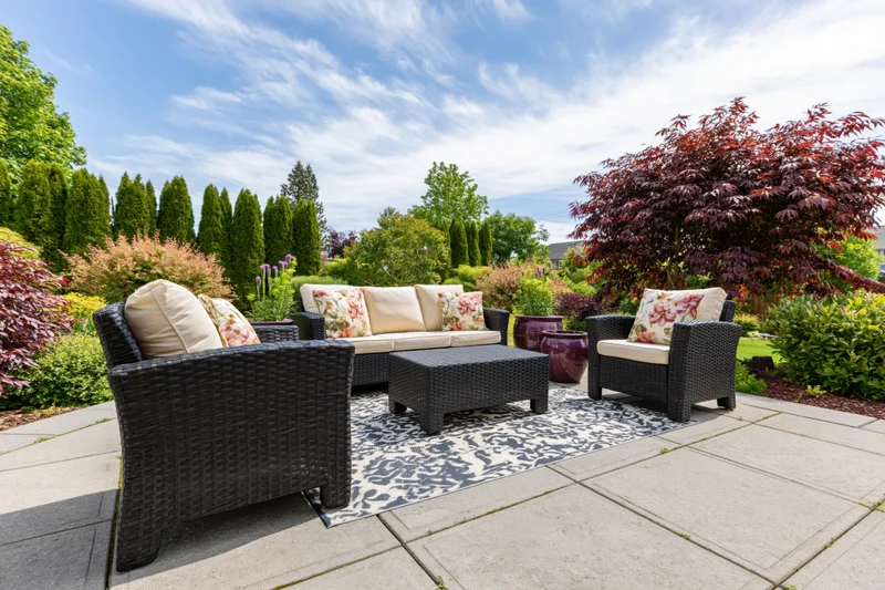 Dark wicker patio furniture set with floral cushions arranged on a stone deck surrounded by landscaped garden with shrubs and trees